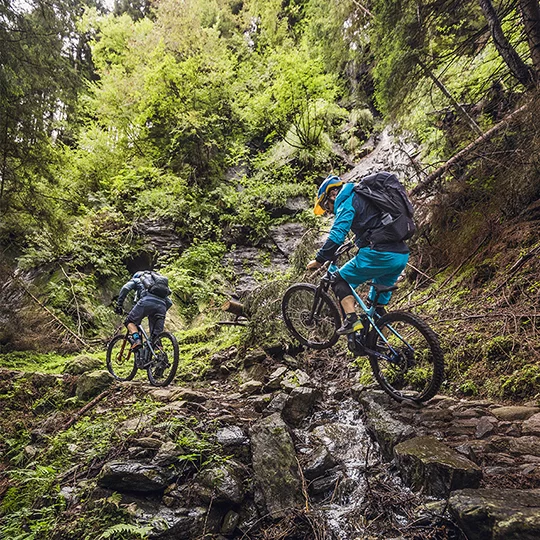 Dos ciclistas de eBike se encuentran en el bosque, uno de ellos salta con su bicicleta sobre un arroyo rodeado de piedras.