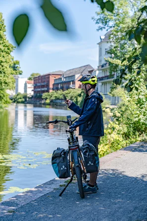 eBike Fahrer steht bei sonnigem Wetter an einem See in der Stadt neben seinem bepackten eBike und bedient sein Smartphone.