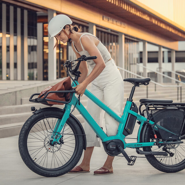 A person in white clothing stands next to a turquoise e-bike with a Bosch motor. The eBike features a front luggage rack with a brown bag placed on it. In the background, there is a modern building with large windows and stairs.