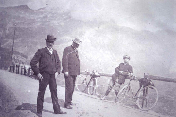 Foto in bianco e nero: due uomini stanno su una strada, mentre un terzo uomo si appoggia a una ringhiera di legno tra le biciclette; sullo sfondo si vedono le montagne.