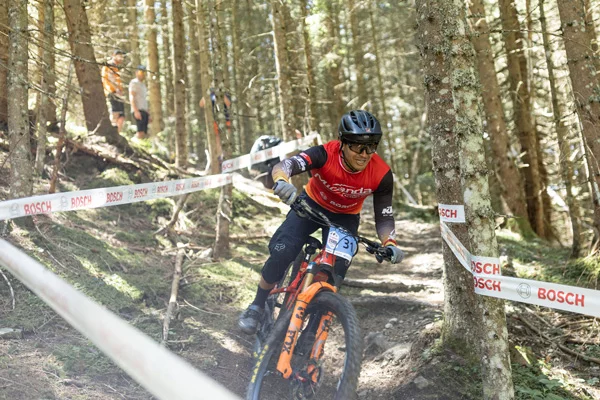 Ein Mann fährt auf einem eMountainbike einen waldigen Downhillpfad entlang dicker Nadelbäume hinunter, der mit Band markiert ist. Zuschauer stehen an der Seite.