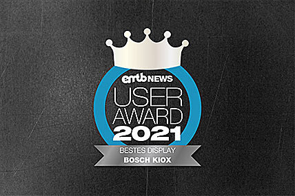 Icon with the text "emtb NEWS USER AWARD 2021": A silver crown above the text, enclosed by a blue circle. Below it says "BEST DISPLAY BOSCH KIOX". The background features a gray concrete design.