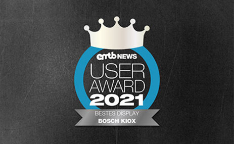 Icon mit dem Text „emtb NEWS USER AWARD 2021“: Eine silberne Krone über der Schrift, umrandet von einem blauen Kreis. Unten steht „BESTES DISPLAY BOSCH KIOX“. Der Hintergrund ist aus grauem Beton.