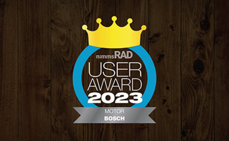 Icon mit dem Text „nimms Rad User Award 2023 Motor Bosch“: Eine goldene Krone über der Schrift, umrandet von einem blauen Kreis. Unten steht „MOTOR BOSCH“. Der Hintergrund ist aus dunklem Holz.