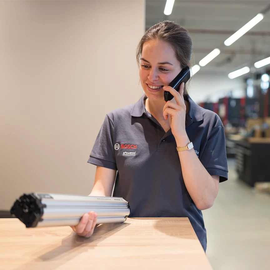A woman wearing a Bosch shirt holds a PowerTube in her hand while talking on the phone. In the background, you can see a workshop environment.