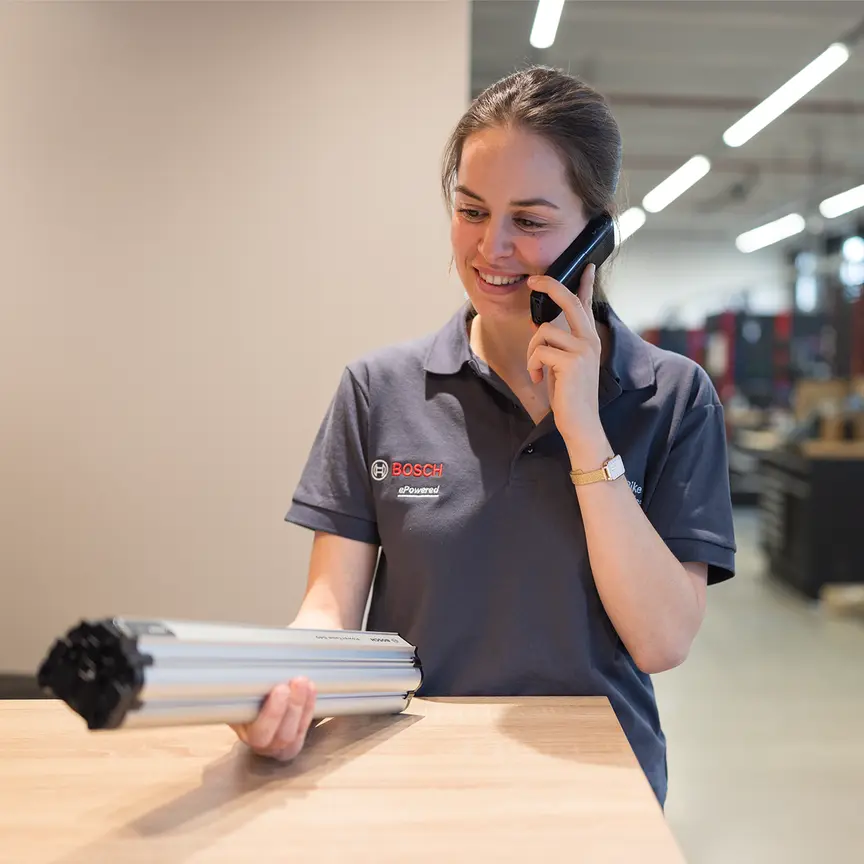 Eine Frau mit einem Bosch-Hemd hält eine PowerTube in der Hand. Gleichzeitig telefoniert sie. Im Hintergrund ist eine Werkstattumgebung zu sehen.