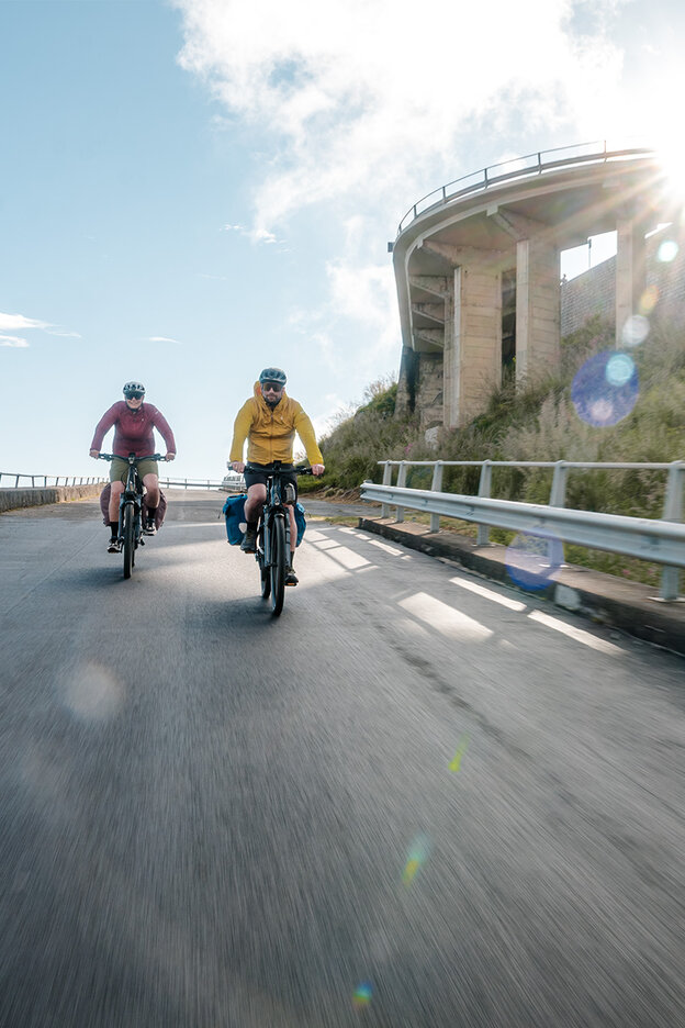 To personer sykler nedover en svingete vei på eBikes. Veien er omkranset av rekkverk og går forbi en steinete klippe. Solen skinner sterkt og skaper lysrefleksjoner i bildet. Scenen gir deg en følelse av eventyr og frihet.