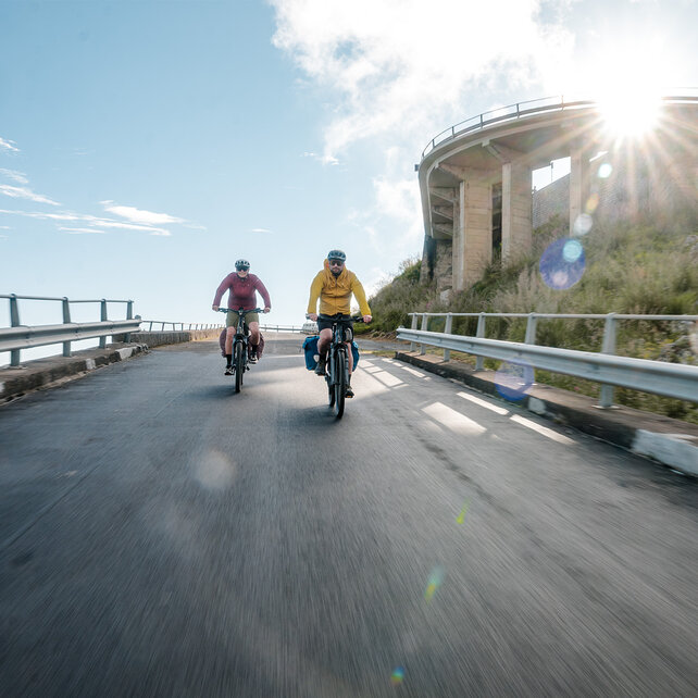 Deux personnes descendent une route sinueuse sur des eBikes. La route est bordée de garde-corps et longe une falaise rocheuse. Le soleil brille et crée des reflets lumineux dans l’image. Cette scène évoque un sentiment d’aventure et de liberté.