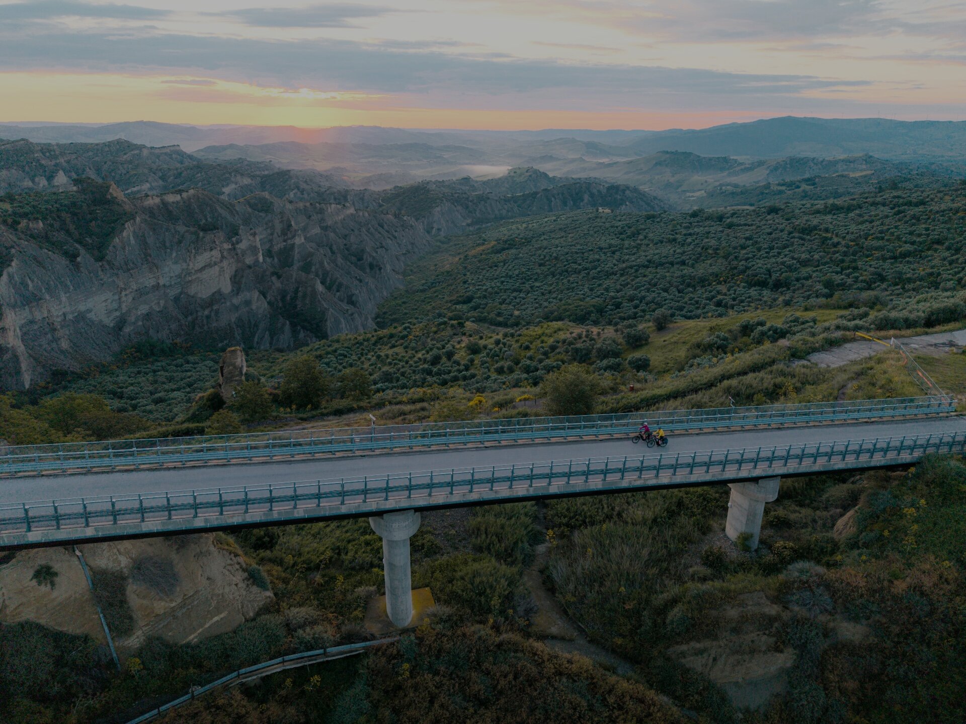 Two people are riding bicycles across a bridge in a mountainous landscape. The area around them is green and hilly, and the setting sun bathes the sky in warm colors.