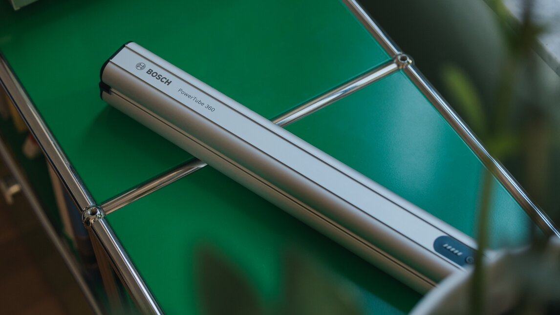 Stylish close-up of a Bosch PowerTube 360 eBike battery lying diagonally on a modern green sideboard with a chrome frame. The battery is showcased like a design object, framed by a blurred plant in the foreground.