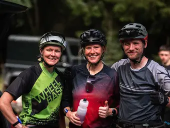 Two women and a man in cycling gear stand together and pose for a photo. Trees are visible in the background.