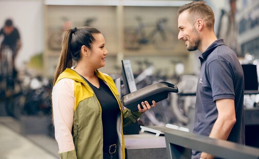 En ung kvinde taler med en kunderådgiver i en cykelbutik. Hun holder et batteri i hånden.