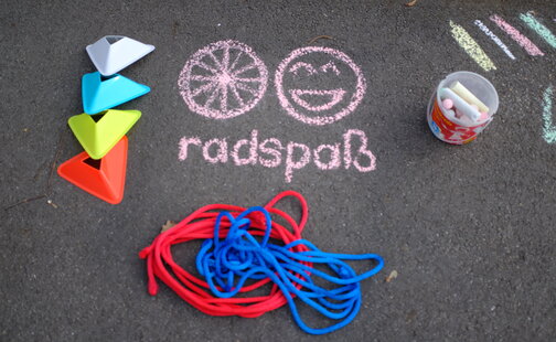 Auf dem Asphalt wurde mit Kreide ein Rad, ein Smiley und das Wort „Radspaß“ gemalt. Daneben liegen eine Kreidekiste, Pylonen und Seile.