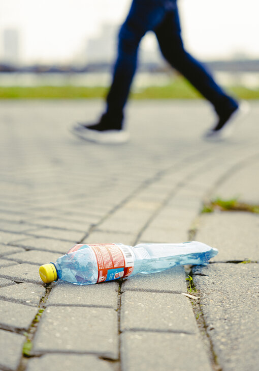 Een plastic fles ligt op de grond, terwijl voeten er vlak achter langs lopen.