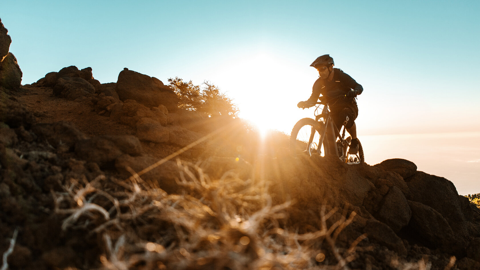 Un(e) pratiquant(e) de eMountainbike monte une colline couverte de pierres au coucher du soleil. Le soleil brille direct dans la caméra et donne l'impression que le(la) eBike pratiquant(e) est une silhouette.