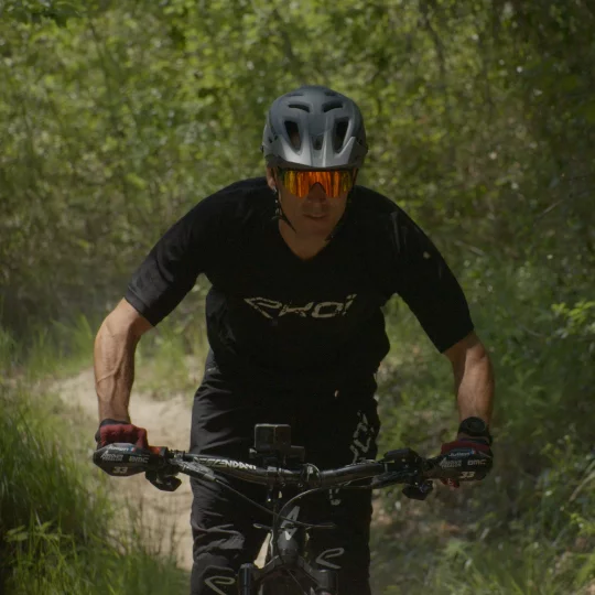 Vue de face d’un homme roulant sur un VTT à assistance électrique sur un parcours très boisé. Il porte un casque et des lunettes de soleil miroir.