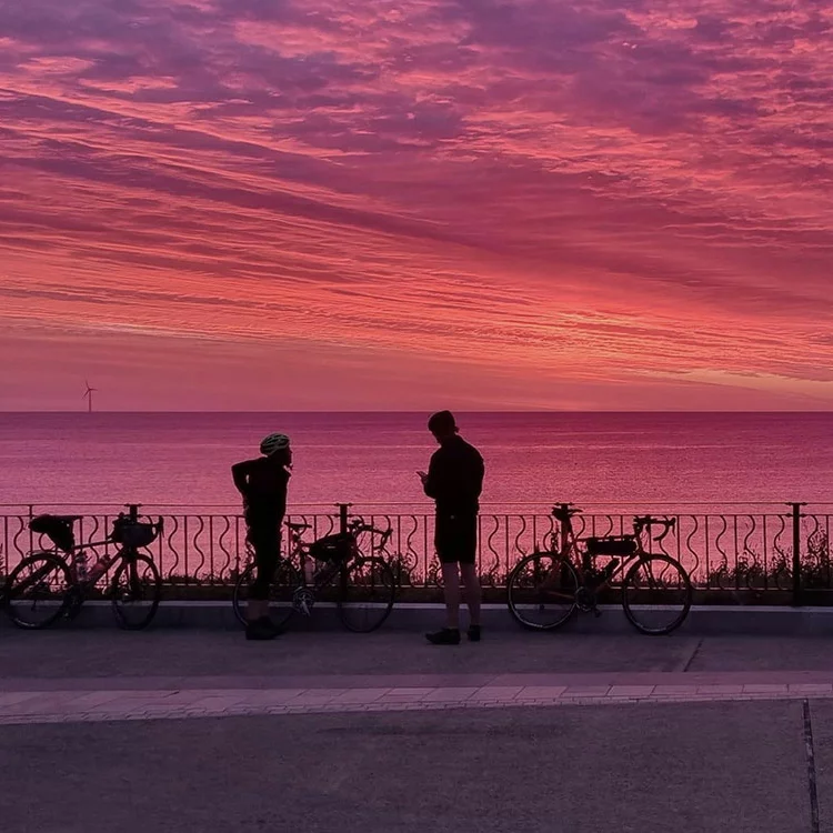 Twee eBike-rijders kijken naar de zonsondergang aan zee, terwijl hun fietsen aan een reling zijn bevestigd.