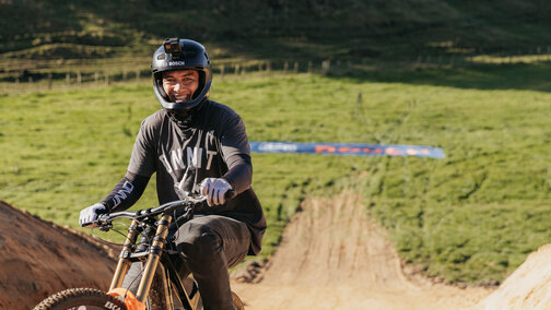 A person wearing a motorcycle helmet sits on an eMountain bike in front of a large jump ramp, smiling at the camera.