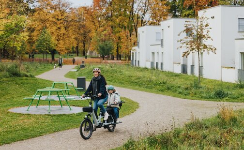Une femme et un enfant, tous deux portant un casque de vélo, roulent sur un Cargo VAE lumineux à travers un parc avec des arbres d’automne et des bâtiments blancs modernes en arrière-plan.