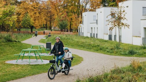 A woman and a child, both wearing bicycle helmets, ride a bright Cargo eBike through a park with autumn trees and modern white buildings in the background.