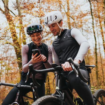 Ein Mann und eine Frau in Fahrradbekleidung und mit Helmen stehen lachend im Herbstwald neben ihren eBikes und schauen gemeinsam auf ein Smartphone.