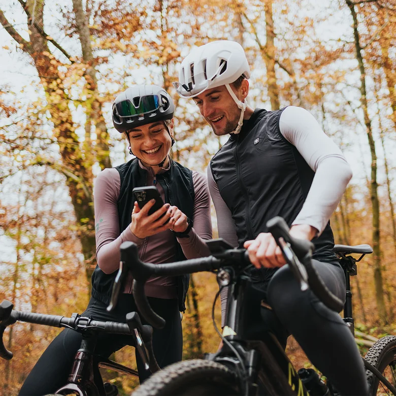 A man and a woman wearing cycling clothing and helmets are laughing together next to their eBikes in an autumn forest, looking at a smartphone.