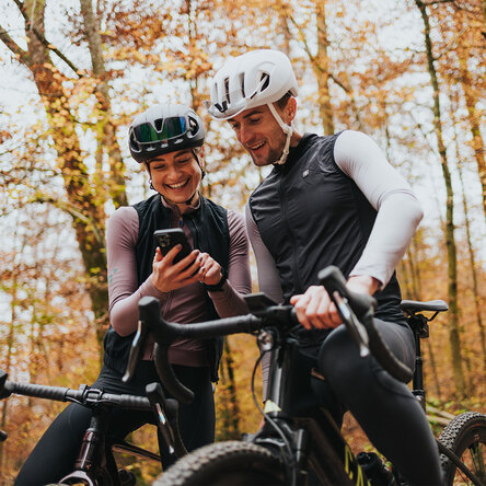 Un uomo e una donna in abbigliamento da ciclista e con il casco ridono accanto alle loro eBike nel bosco autunnale e guardano insieme uno smartphone.