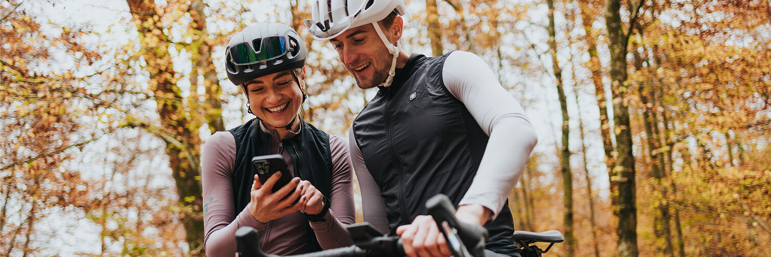 A man and a woman wearing cycling clothing and helmets are laughing together next to their eBikes in an autumn forest, looking at a smartphone.