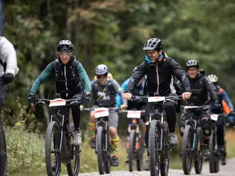 A group of cyclists in gear with race numbers on their handlebars is visible. Trees are in the background.