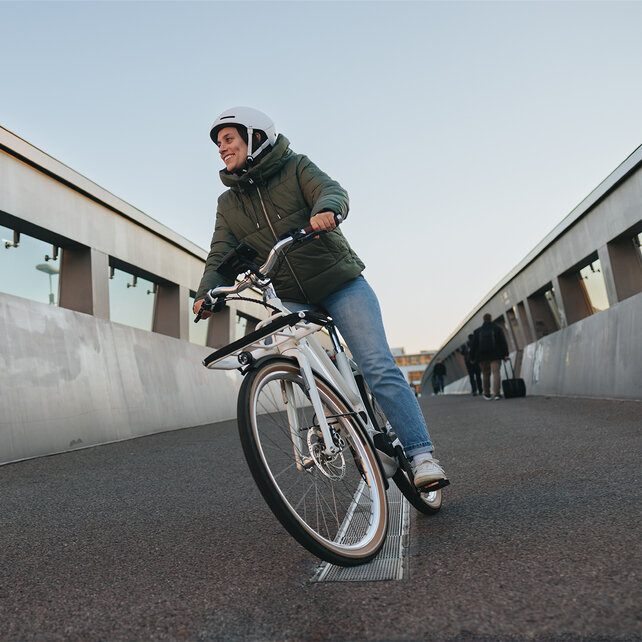 Woman wearing a helmet and winter jacket rides her white eBike over a bridge.