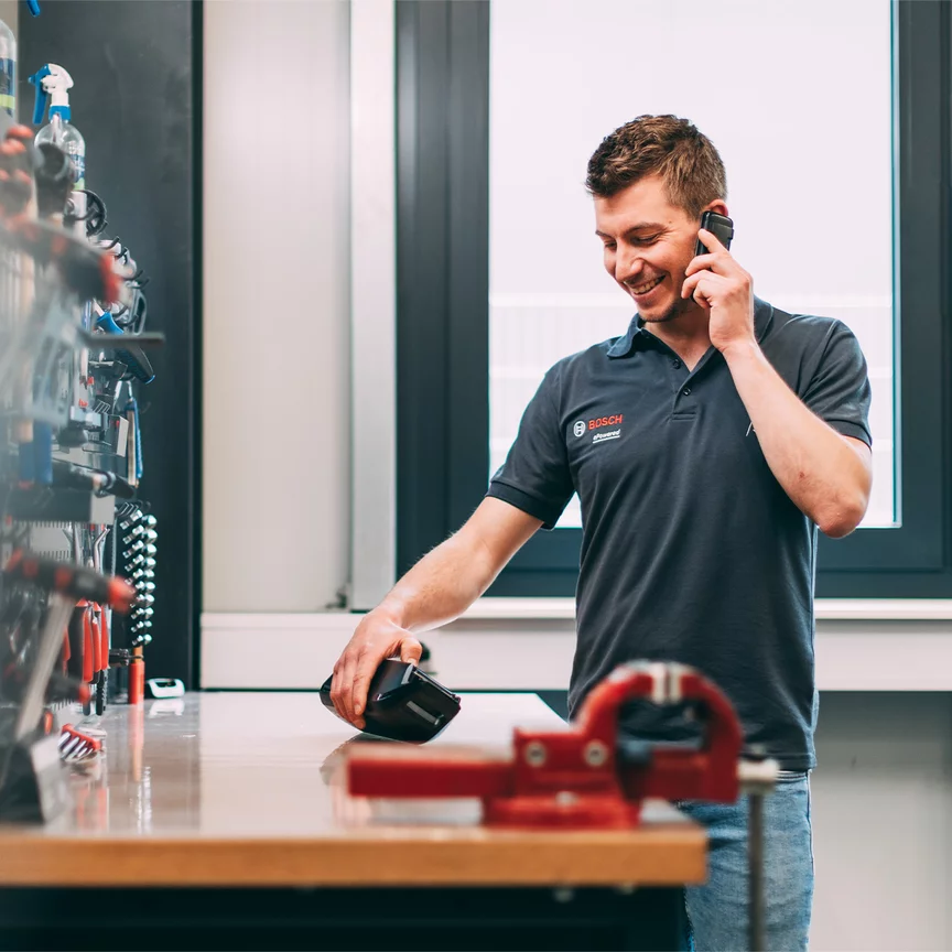 Ein Bosch-Mitarbeiter telefoniert mit einem Lächeln im Gesicht, während er auf ein Bosch-Produkt schaut, das auf einem Werktisch liegt.