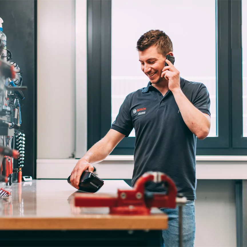 A Bosch employee is smiling as he talks on the phone, while looking at a Bosch product on a workbench.