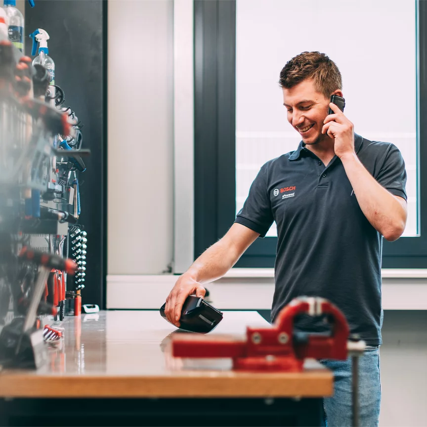 Ein Bosch-Mitarbeiter telefoniert mit einem Lächeln im Gesicht, während er auf ein Bosch-Produkt schaut, das auf einem Werktisch liegt.