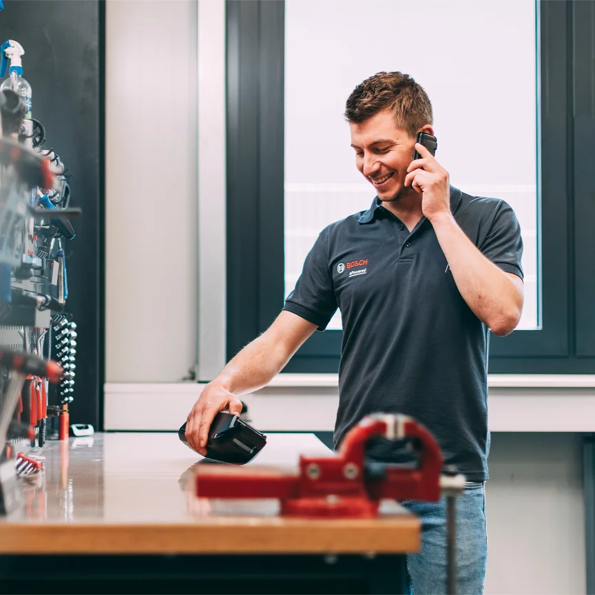 A Bosch employee is smiling as he talks on the phone, while looking at a Bosch product on a workbench.