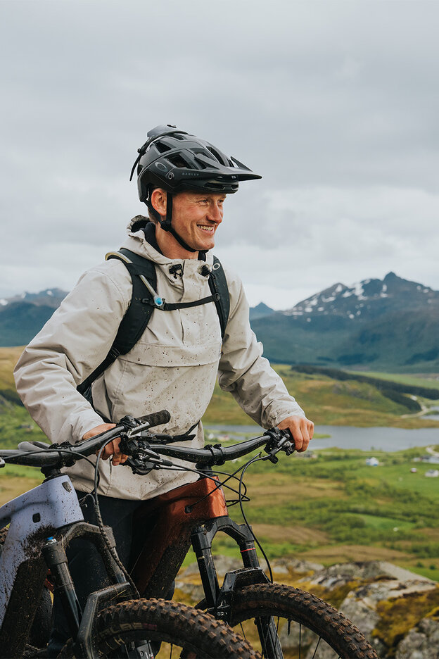 Zwei Personen stehen mit E-Mountainbikes auf einem Hügel und blicken in die Ferne. Sie tragen Helme und Outdoor-Kleidung. Im Hintergrund ist eine grüne Landschaft mit Bergen und einem See zu sehen. Es ist bewölkt.