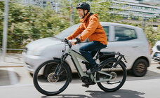 A man is driving an eBike on a road, with parked cars, bushes, and buildings visible in the background.
