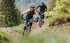 Two eMountain bike riders ride down an earthy path between bushes, in the background you can see mountains.