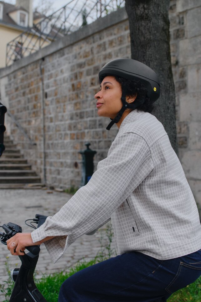 Una persona con casco espera en su eBike en un semáforo peatonal en rojo. Levanta ligeramente la vista mientras está en una calle empedrada junto a un viejo muro de piedra y un semáforo histórico.