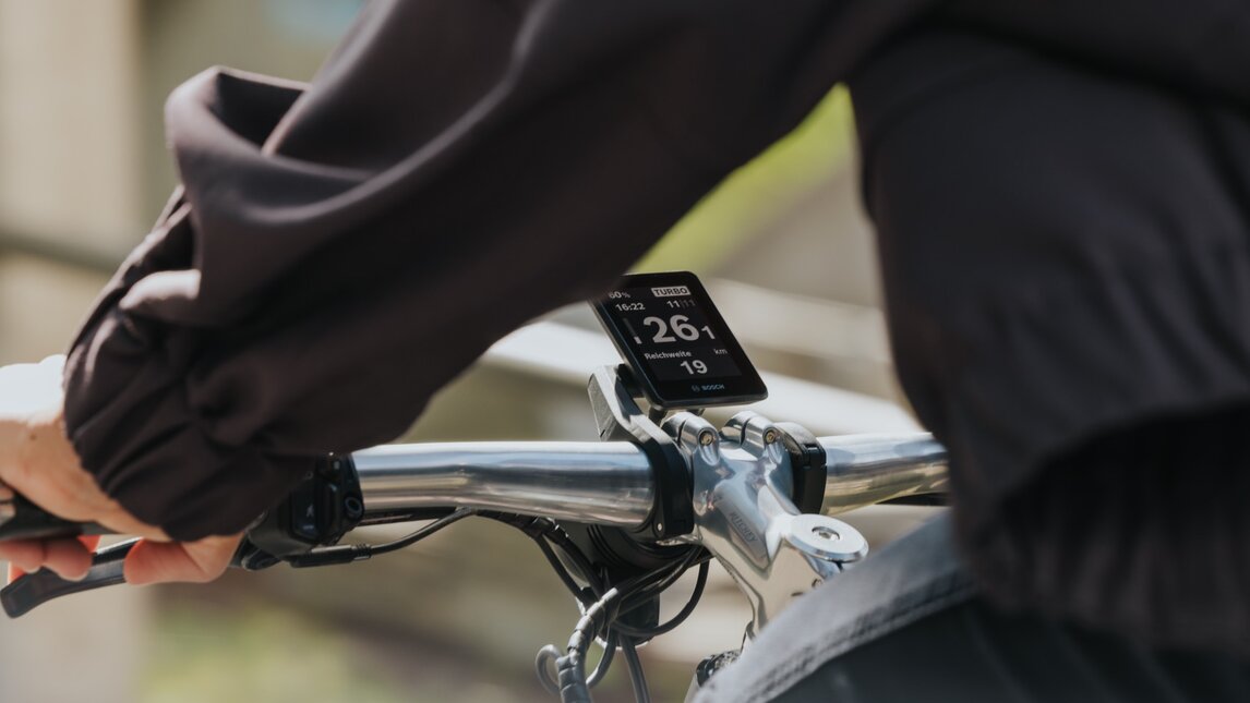 Rider's perspective of the cockpit on an eBike while riding. The focus is on the Bosch Intuvia display 200 on the handlebars, showing the current speed and range in Turbo mode. The background is blurred from the motion.