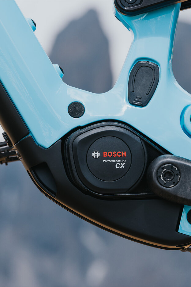 Nærbilde av en elsykkelmotor fra Bosch Performance Line CX. Sykkelen har en lyseblå rammefarge og er plassert mot en uskarp bakgrunn med fjell.