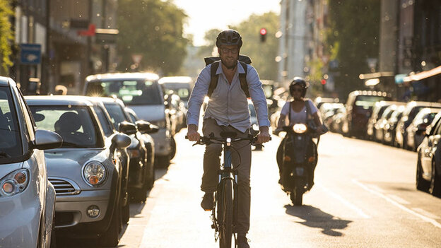 eBike rider is riding along a road, with a motorcyclist visible behind and cars parked to his left on the street.