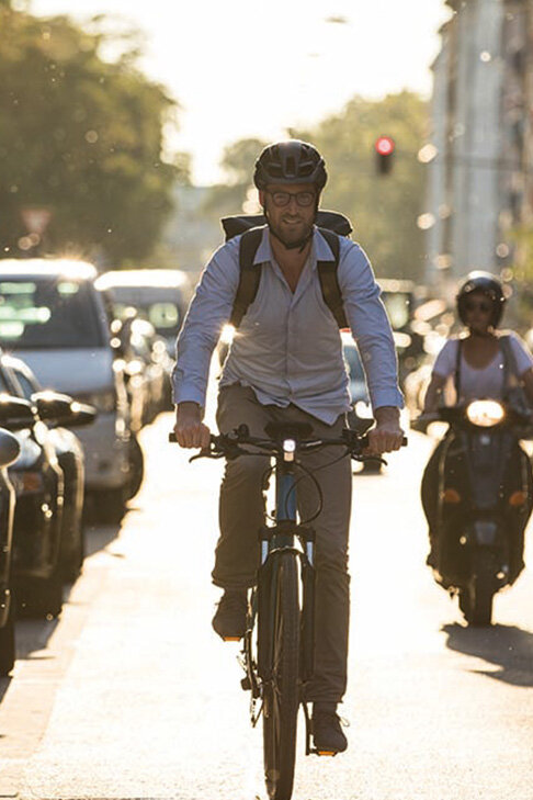 eBike rider is riding along a road, with a motorcyclist visible behind and cars parked to his left on the street.