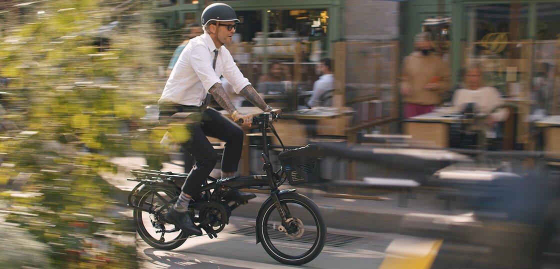 Bob Harper rides past a café on an eBike, where you can see blurred people sitting outside at tables.