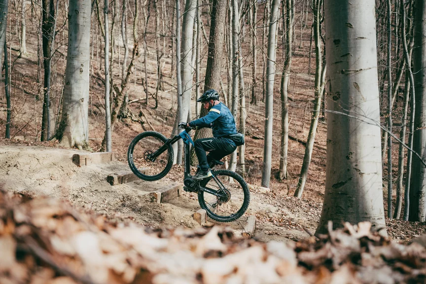Ein Mann fährt auf einem eMountainbike im Wald aus Holz gebauteTreppenstufen hinauf. Die Bäume im Wald sind kahl und es liegt Laub auf dem Boden.