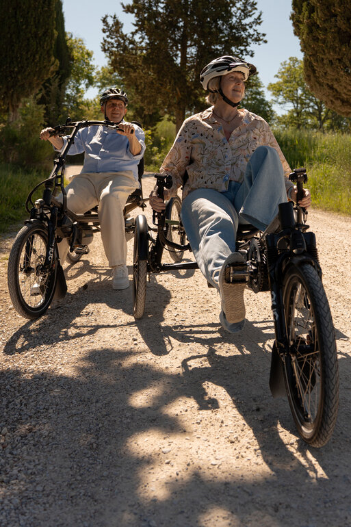 Deux personnes roulent côte à côte sur des eBikes spéciaux à trois roues sur un chemin de gravier à la campagne.