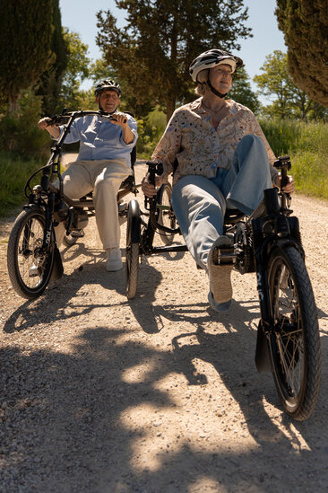 Deux personnes roulent côte à côte sur des eBikes spéciaux à trois roues sur un chemin de gravier à la campagne.
