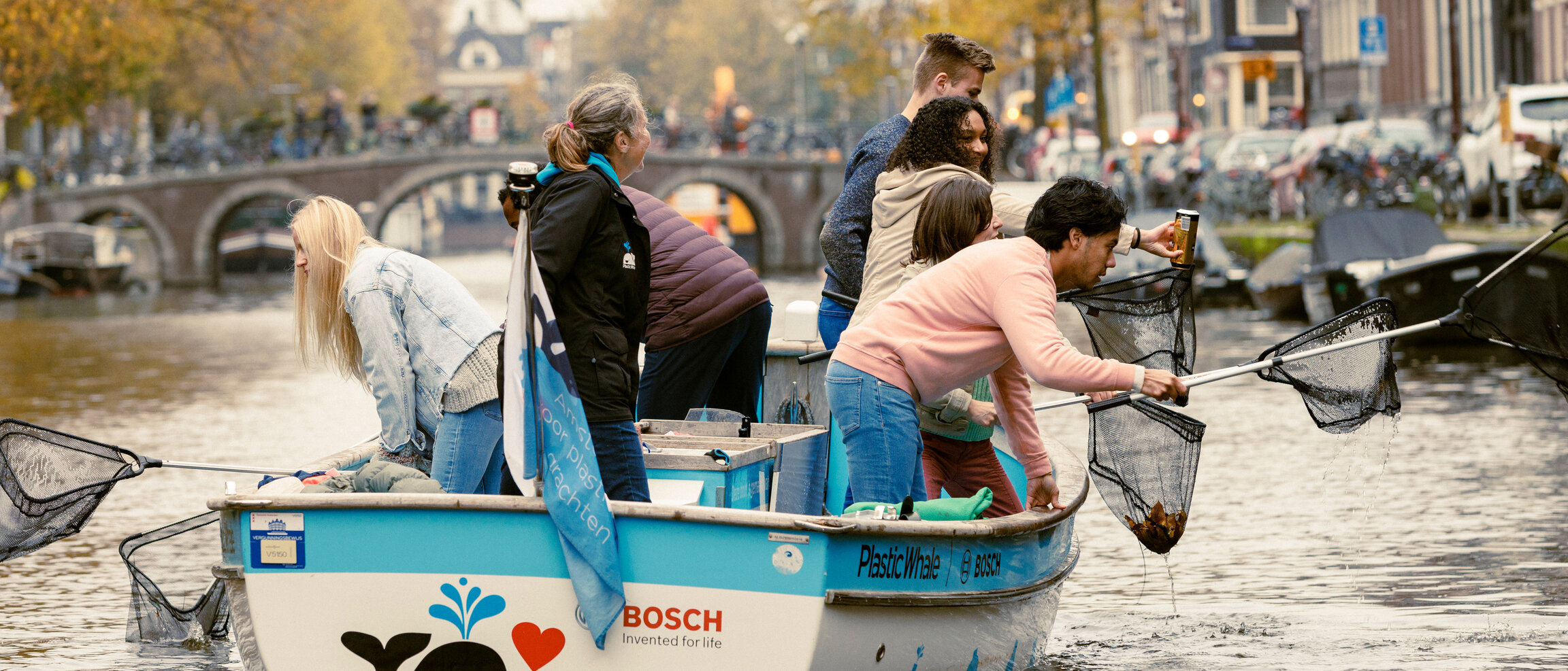 Meerdere mensen zitten op een bootje op een gracht en vissen met schepnetten dingen uit het water.
