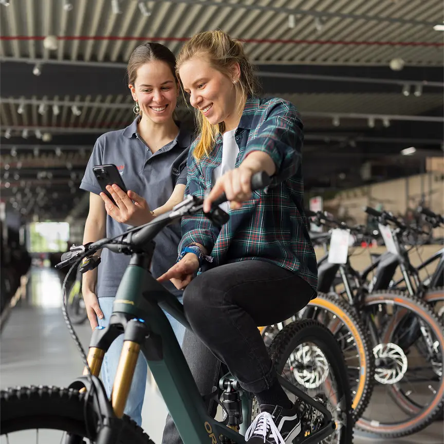Zwei Personen stehen in einem Fahrradgeschäft. Eine Person sitzt auf einem grünen E-Bike und trägt eine karierte Jacke sowie schwarze Jeans und schwarze Schuhe. Die andere Person steht daneben und hält ein Smartphone. Im Hintergrund sind mehrere Fahrräder und eine moderne Einrichtung zu sehen.