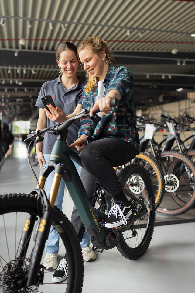 Two people are in a bike shop. One person is sitting on a green eBike, wearing a plaid jacket, black jeans, and black shoes. The other person stands next to them, holding a smartphone. In the background, you can see several bikes and modern furnishings.
