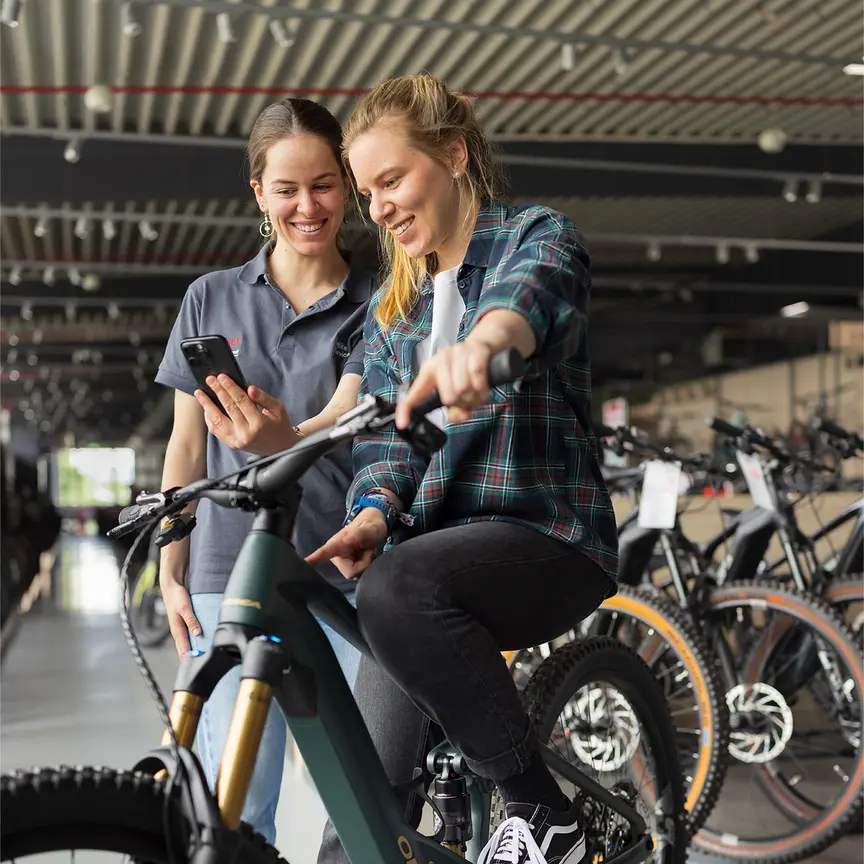 Two people are in a bike shop. One person is sitting on a green eBike, wearing a plaid jacket, black jeans, and black shoes. The other person stands next to them, holding a smartphone. In the background, you can see several bikes and modern furnishings.